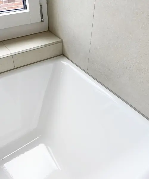 Das Bild zeigt eine neue, glänzende Badewanne in einem modernen Badezimmer mit hellen Fliesen und einer Fensterbank aus gefliesten Elementen. Die Wandfliesen sind in einem neutralen Beige-Ton gehalten, was eine elegante und minimalistische Optik schafft. Die frisch verfugten Silikonfugen zwischen der Badewanne und den Fliesen sorgen für eine wasserdichte Abdichtung, die das Eindringen von Feuchtigkeit verhindert und die Langlebigkeit der Installation erhöht. Eine fachgerechte Silikonversiegelung ist entscheidend, um Schimmelbildung und Feuchtigkeitsschäden zu vermeiden, insbesondere in Feuchträumen wie Badezimmern und Duschen. Neu versiegelte Fugen bieten nicht nur Schutz, sondern tragen auch zu einem sauberen und gepflegten Erscheinungsbild bei.