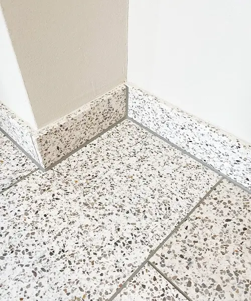 Das Bild zeigt eine Raumecke mit neu verlegten Terrazzo-Fliesen auf dem Boden und an der Sockelleiste. Die grau-weißen Terrazzo-Fliesen haben ein modernes, klassisches Design mit kleinen farbigen Gesteinseinschlüssen, das typisch für hochwertige Bodenbeläge in Innenräumen ist. Die Fugenlinien sind sauber und gleichmäßig, was auf eine fachgerechte Verlegung und Versiegelung hinweist. Terrazzo ist ein robuster und langlebiger Bodenbelag, der oft in Badezimmern, Küchen und Eingangsbereichen verwendet wird. Die saubere Fugenverarbeitung sorgt für eine wasserdichte und pflegeleichte Oberfläche, die sich leicht reinigen lässt und resistent gegen Feuchtigkeit ist.