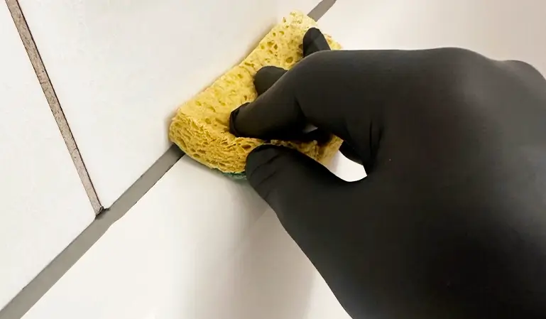 Hand mit schwarzem Handschuh reinigt Silikonfugen zwischen weißen Fliesen mit einem gelben Reinigungsschwamm