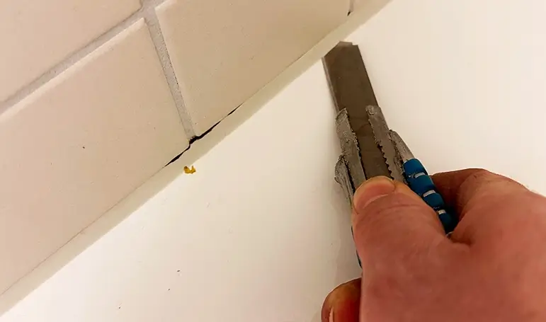 Das Bild zeigt eine Hand mit einem schwarzen Einweghandschuh, die mit einem gelben Reinigungsschwamm eine verschmutzte Silikonfuge zwischen weißen Fliesen reinigt. Die Fuge war mit Schmutz, Seifenresten und Ablagerungen bedeckt, doch nach der Reinigung erscheint sie sauber und hygienisch. Diese Technik entfernt Schimmel, Bakterien und Verfärbungen, sodass die Silikonfuge länger hält und nicht beschädigt wird.